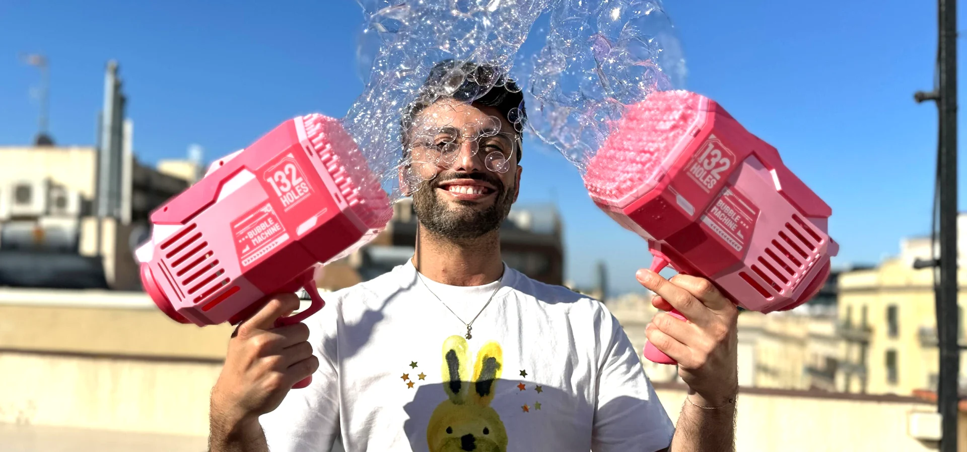 Dimitris smiling on a rooftop under a blue sky, bubbles floating above his head, holding two pink bubble guns