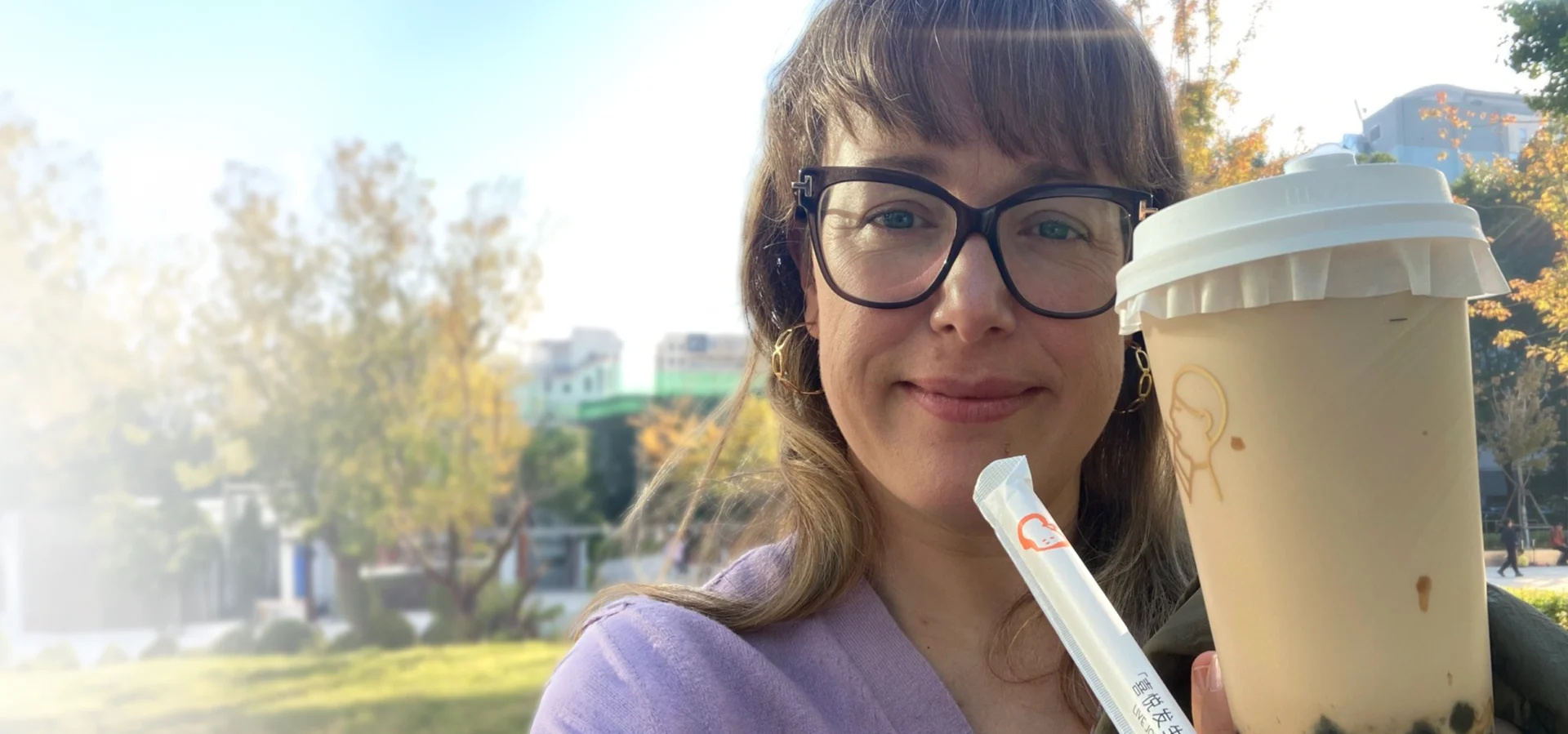 FunPlus 'Meet Eleanor' banner variant, without text overlay: a woman in glasses smiles outdoors, holding a milk tea cup with a straw, with autumn trees and buildings in the background.